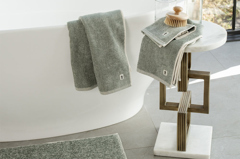 A set of gray bath towels and a matching bath mat styled in a modern bathroom setting next to a white freestanding bathtub and a decorative side table.