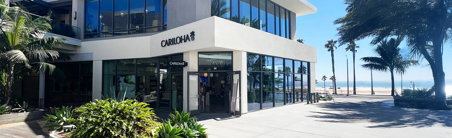 The exterior of the Cariloha store, featuring a modern white facade, large glass windows, and a beachfront setting with palm trees and ocean view under a clear blue sky.