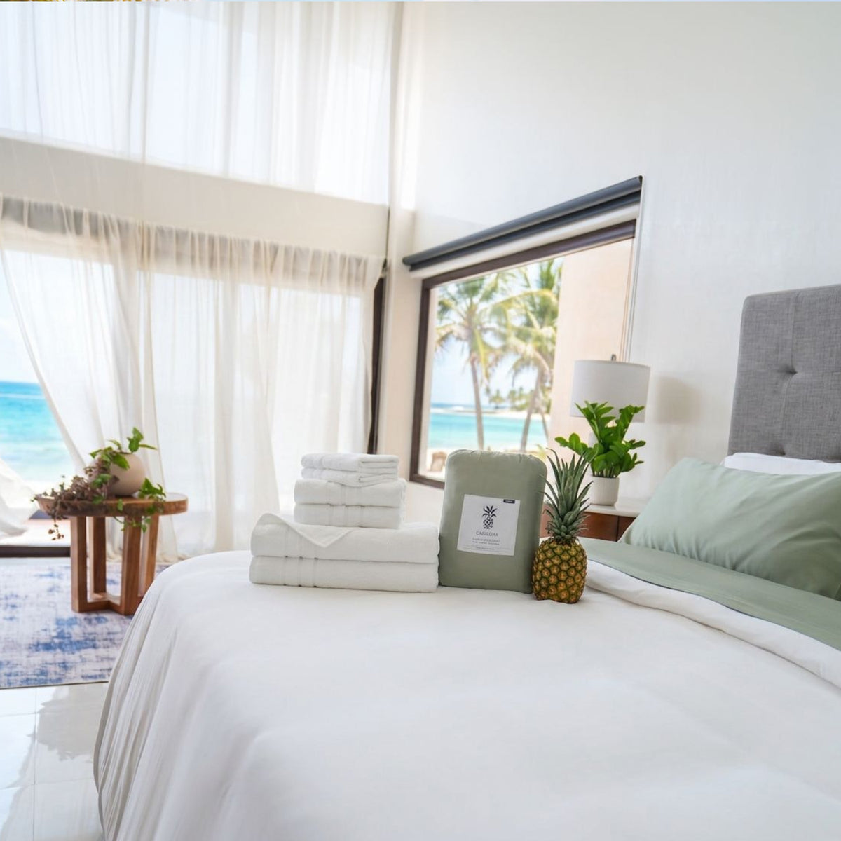 A neatly made bed in a bright, modern bedroom with white linens, pale green pillows and a folded towel, plus a small pineapple  and a package of bamboo sheets on the bed. Large windows show an ocean view with palm trees, and sheer white curtains frame the left window.