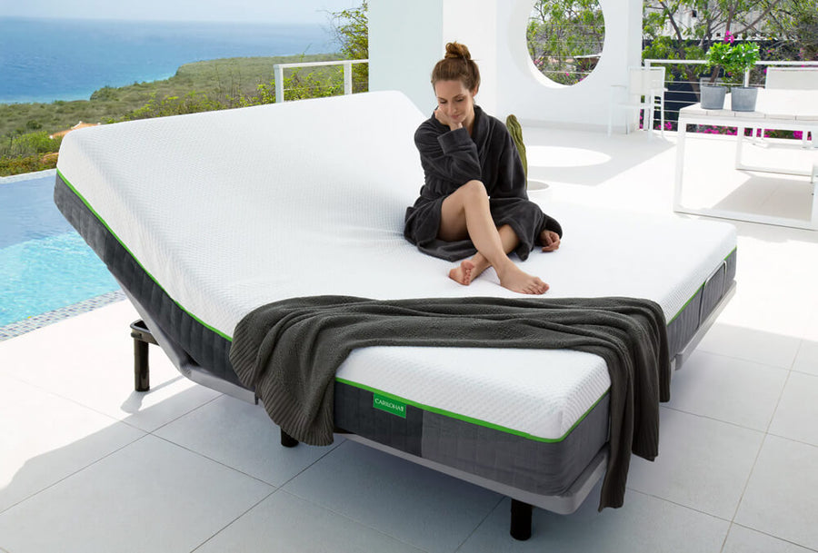A woman in a dark robe sits on a white adjustable bed with gray side panels and green accents, set in a bright modern outdoor patio area with a pool and ocean view.