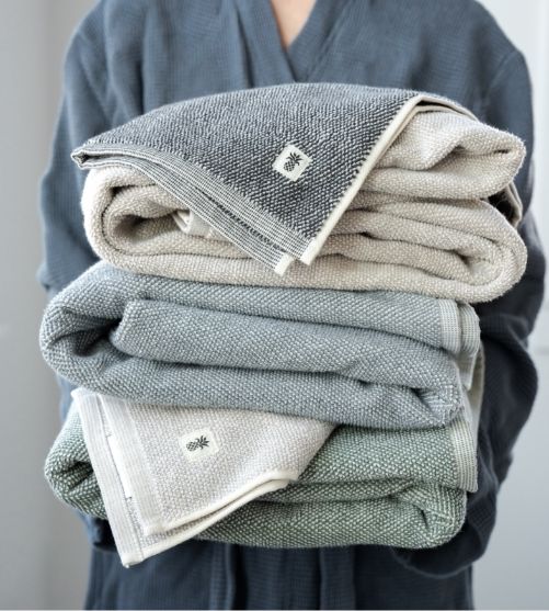 Person in a Cariloha bamboo robe holding a stack of folded bath towels in grey, sage, and stone colors