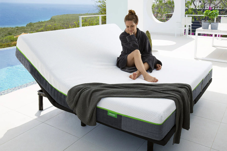 A woman sits on a white adjustable mattress on a bed frame in a bright, modern outdoor patio setting with a pool and ocean view. The Cariloha classic mattress has a white top, dark gray sides, and a thin green accent line.