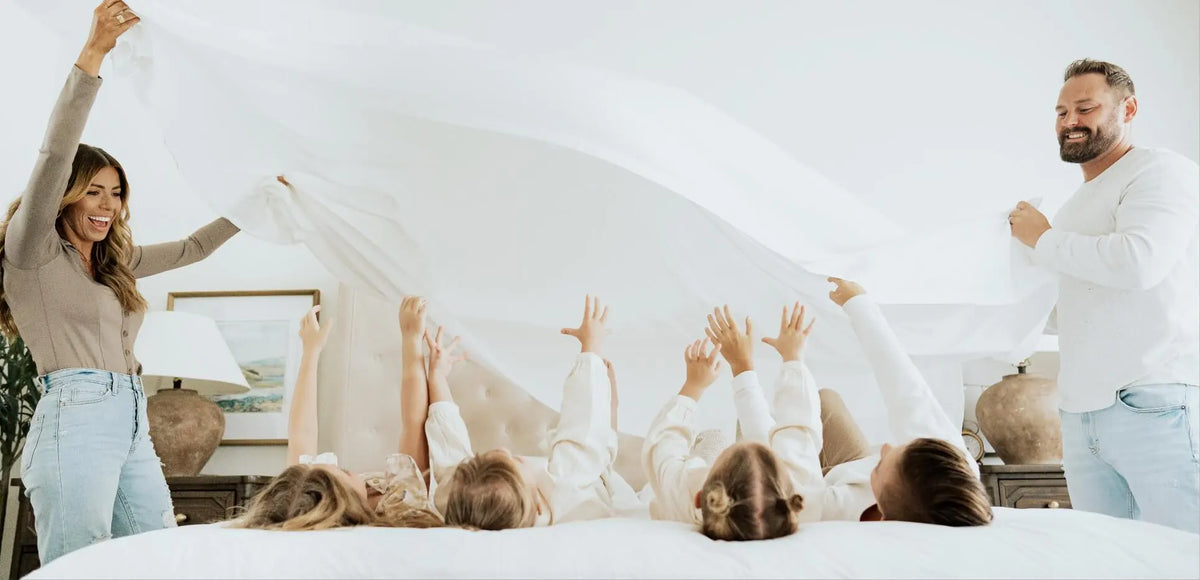 A family of five playing on a bed with a large white sheet, with two adults holding the sheet up on either side. The children are lying on their backs with their hands reaching up into the white fabric.
