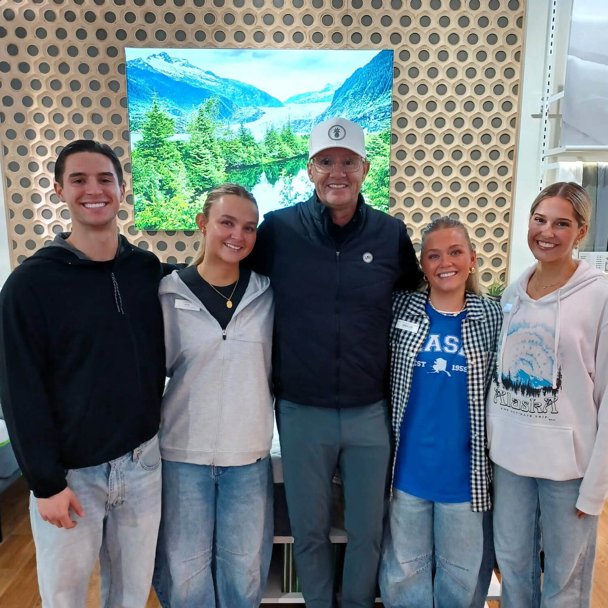 Five people standing together in a retail store interior, with a large framed mountain-and-forest landscape print on the wall behind them. The center person is wearing a white cap, dark jacket, and gray pants, while the others wear casual clothing.