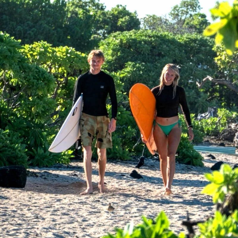 A man and a woman walking on a sandy beach carrying surfboards, with lush green tropical foliage in the background.