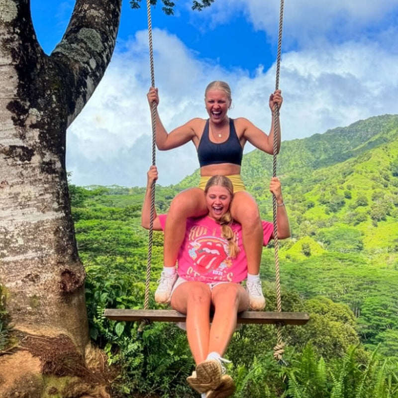 Two women posing together on a rope swing suspended from a tree, with a lush green mountain landscape and blue sky in the background. One woman wears a black sports bra and yellow shorts, while the other wears a pink Rolling Stones graphic T-shirt and white sneakers.