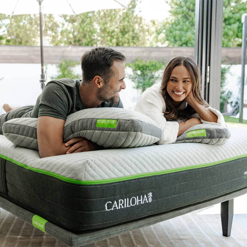 A man and a woman lie on a mattress outdoors on a patio, each holding a gray pillow with a green label. The mattress has a dark gray side panel with the brand name CARILOHA visible and a bright green stripe detail.