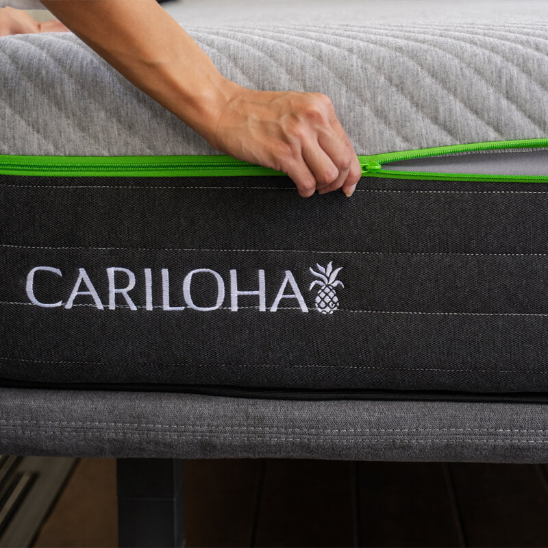 A close-up of a person's hand unzipping a mattress, showing the brand name CARILOHA embroidered in white on a dark gray fabric panel with a bright green zipper.