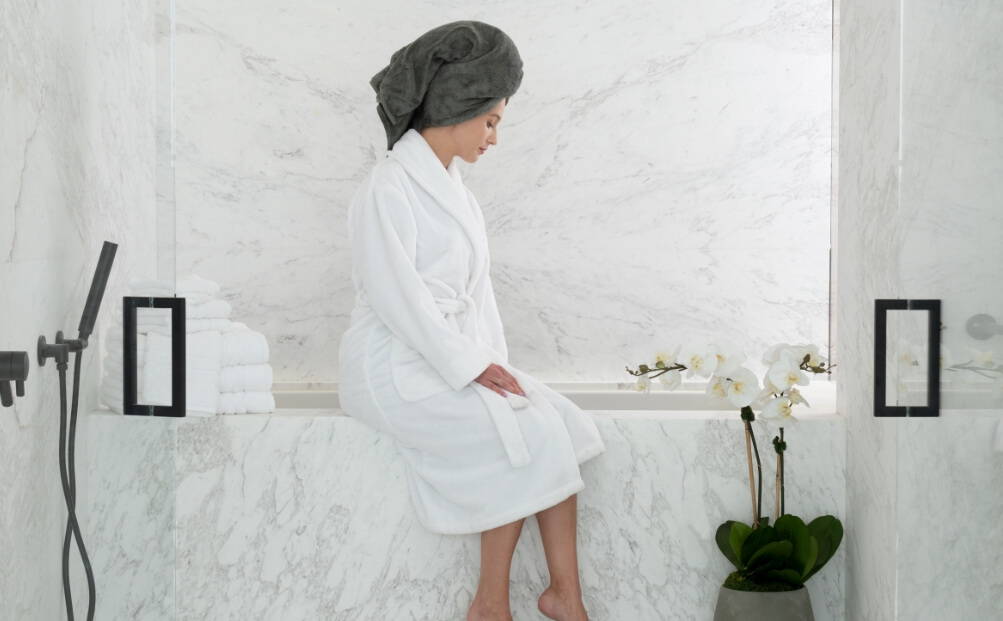 Woman in a Cariloha bamboo bathrobe with a towel wrapped around her hair, seated on the edge of a marble soaking tub beside folded towels and orchids