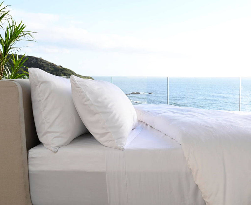 White Cariloha bamboo-viscose bed linens styled on an outdoor bed overlooking the ocean, with a potted palm alongside
